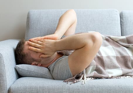man laying on couch with hands over face and blanket on