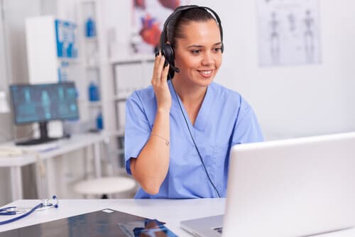 Woman in blue medical scrubs sitting in front of a computer and talking on the phone via headset.